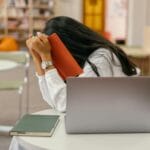 Kurs prawa jazdy dla nieśmiałych: jak przełamać blokadę, zadawać pytania i prosić o trudniejsze trasy Zestresowana kursantka chowa twarz w książce przy laptopie w bibliotece