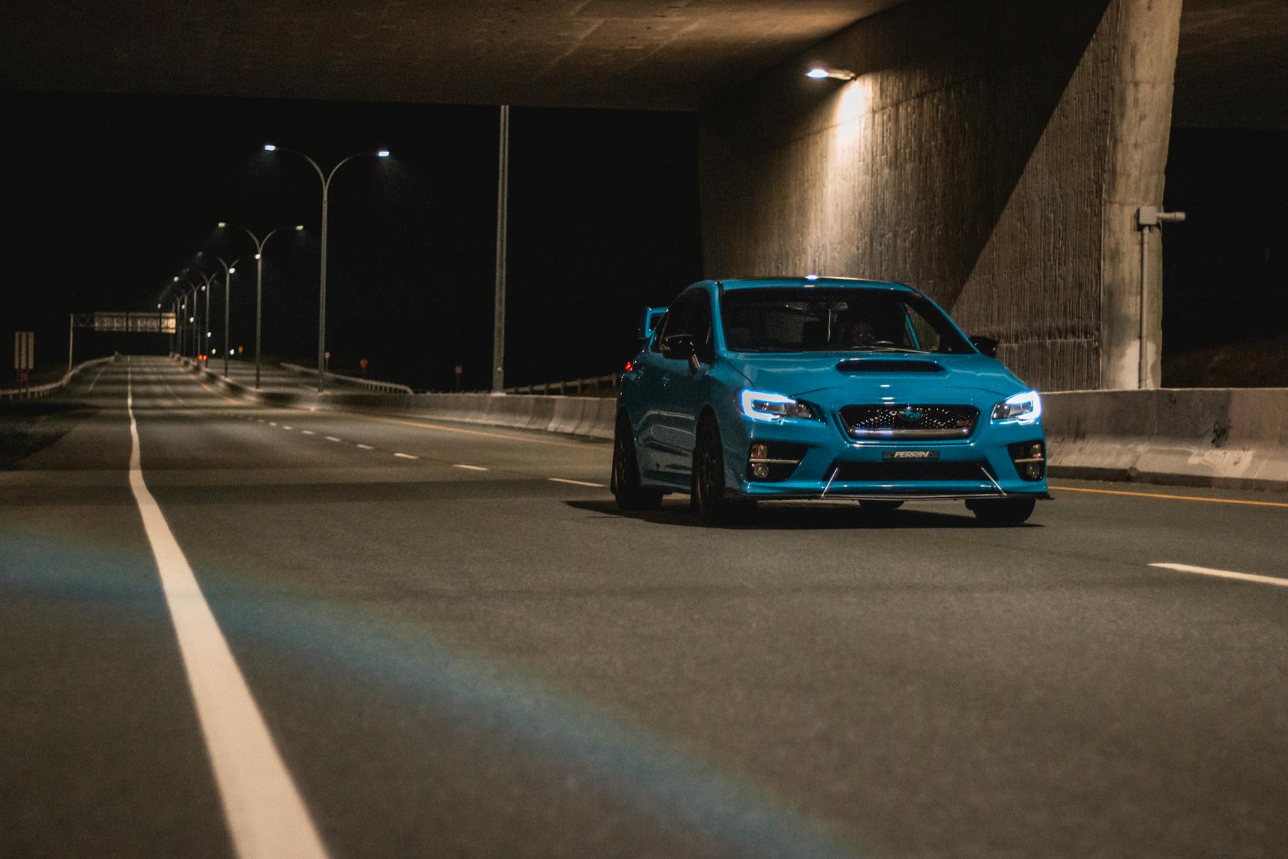 Niebieskie auto z włączonymi światłami jedzie nocą po oświetlonej drodze